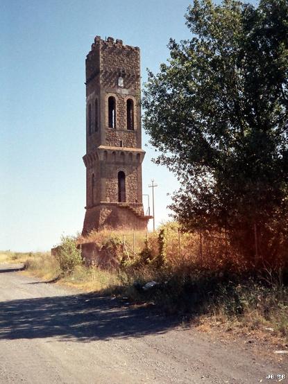 Farbfoto des Turms eines ehemaligen Wasserhebewerks in der Nähe von Pomezia, Lazio, Italien. Im Vordergrund rechts ein grüner Baum, daran anschließend Gräser, links eine Schotterstraße mit Schatten, im Hintergrund blassblauer Himmel. Olympus 35RC auf Orwo 200-Film.