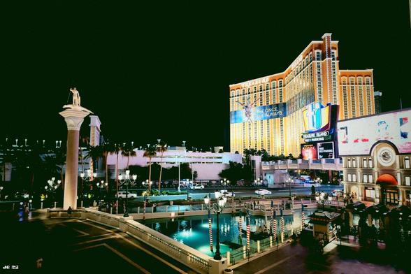 Nachtaufnahme aus Las Vegas, Nevada, USA. Hier ist bei tiefschwarzer Nacht viel Licht vorhanden, zum Beispiel durch das hell angestrahlte gelbe Kasino (Treasure Island) rechts von der Bildmitte. Im Vordergrund türkisfarben ein Teil einer Venedig nachempfundenen künstlichen Lagune.