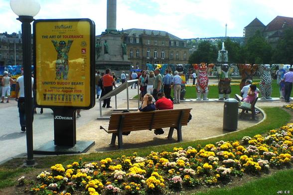 Farbfoto, aufgenommen während der Ausstellung "United Buddy Bears (The Art of Tolerance)" auf dem Schlossplatz in Stuttgart 2008. Links eine Bildsäule mit Info zu der Installation. In Bildmitte viele Besucher und rechts etliche der aufgestellten Bären. Im Hintergrund die Jubiläumssäule und das Schloss und blassblauer Himmel mit weißen Wolken.