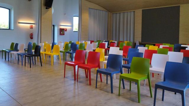 Blick in das Auditorium eines Kunstmuseums, in dem schön geordnet Kunststoffstühle in mehreren Farben - blau, grün, rot, gelb, weiß, schwarz - in Reih und Glied aufgestellt sind.