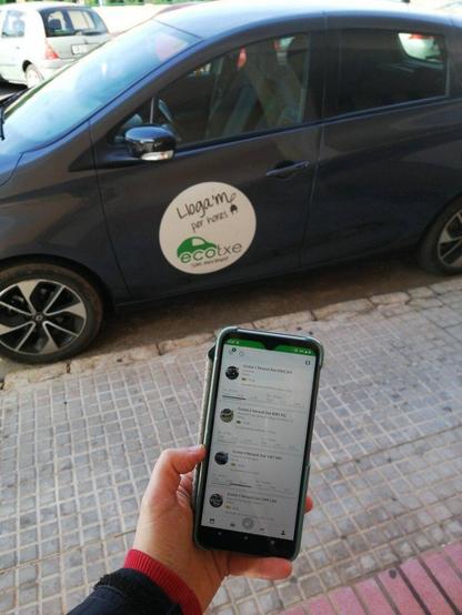 Una mà sosté un mòbil amb l'aplicació d'Ecotxe oberta. En segon pla, es veu un Ecotxe de perfil aparcat al carrer amb un cartell a la porta que diu "Lloga'm per hores".