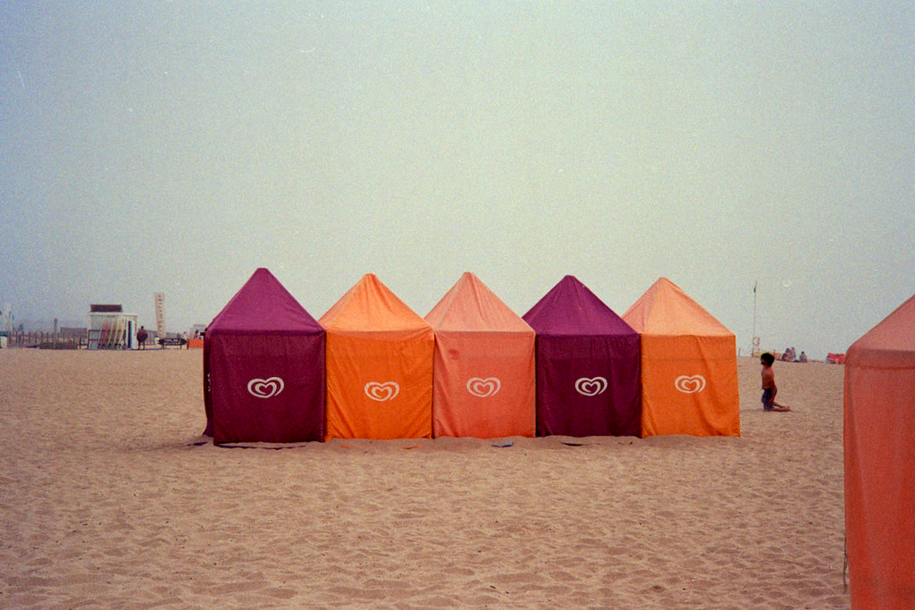 Una serie de tiendas para cambiarse de ropa en la playa con colores morados y naranjas. Un hombre de rodillas en un lado. Niebla en el cielo.